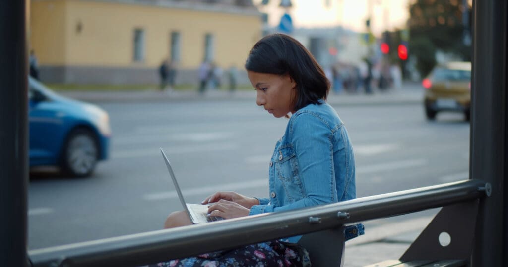 Back view of woman working on laptop sitting on bus stop in city. american young female using computer sitting on bench on city street